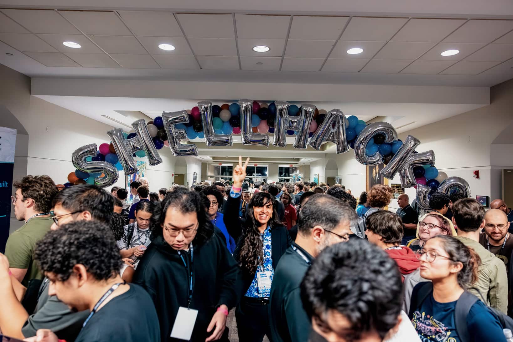 background image of shellhacks, the biggest hackathon in Florida, displaying a sea of software enginers networking and a young individual in the middle of it, smiling and making a peace sign in the middle of the crowd
